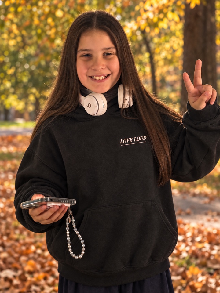 Layla smiling outdoors in autumn, wearing her Love Loud hoodie and headphones, holding her phone and making a peace sign