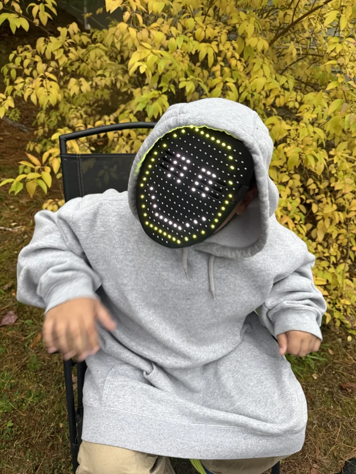 Rocco seated beside golden autumn leaves, wearing a gray hoodie and a light-up mask with a different face
